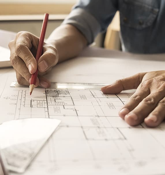 A hand holding a pencil over building plans. 