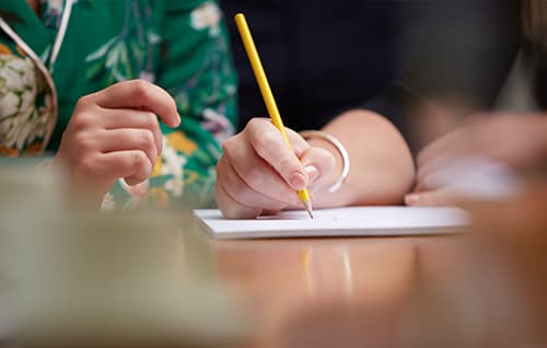 Two people working. One is writing with a yellow pencil. 